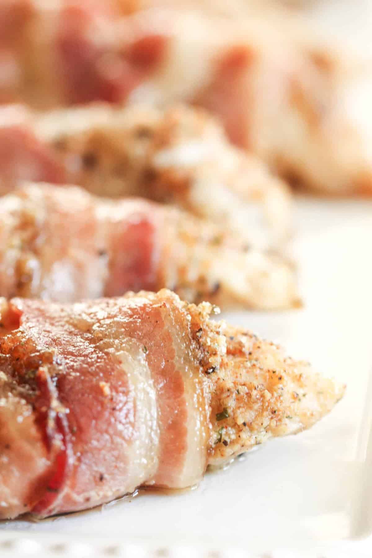 Close-up of seasoned chicken breasts wrapped in bacon, arranged on a white plate, with the focus on the texture of the meat and seasoning.