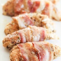 Close-up of several bacon-wrapped chicken pieces seasoned with spices, arranged in a row on a white plate.