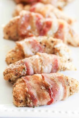 Close-up of several bacon-wrapped chicken pieces seasoned with spices, arranged in a row on a white plate.