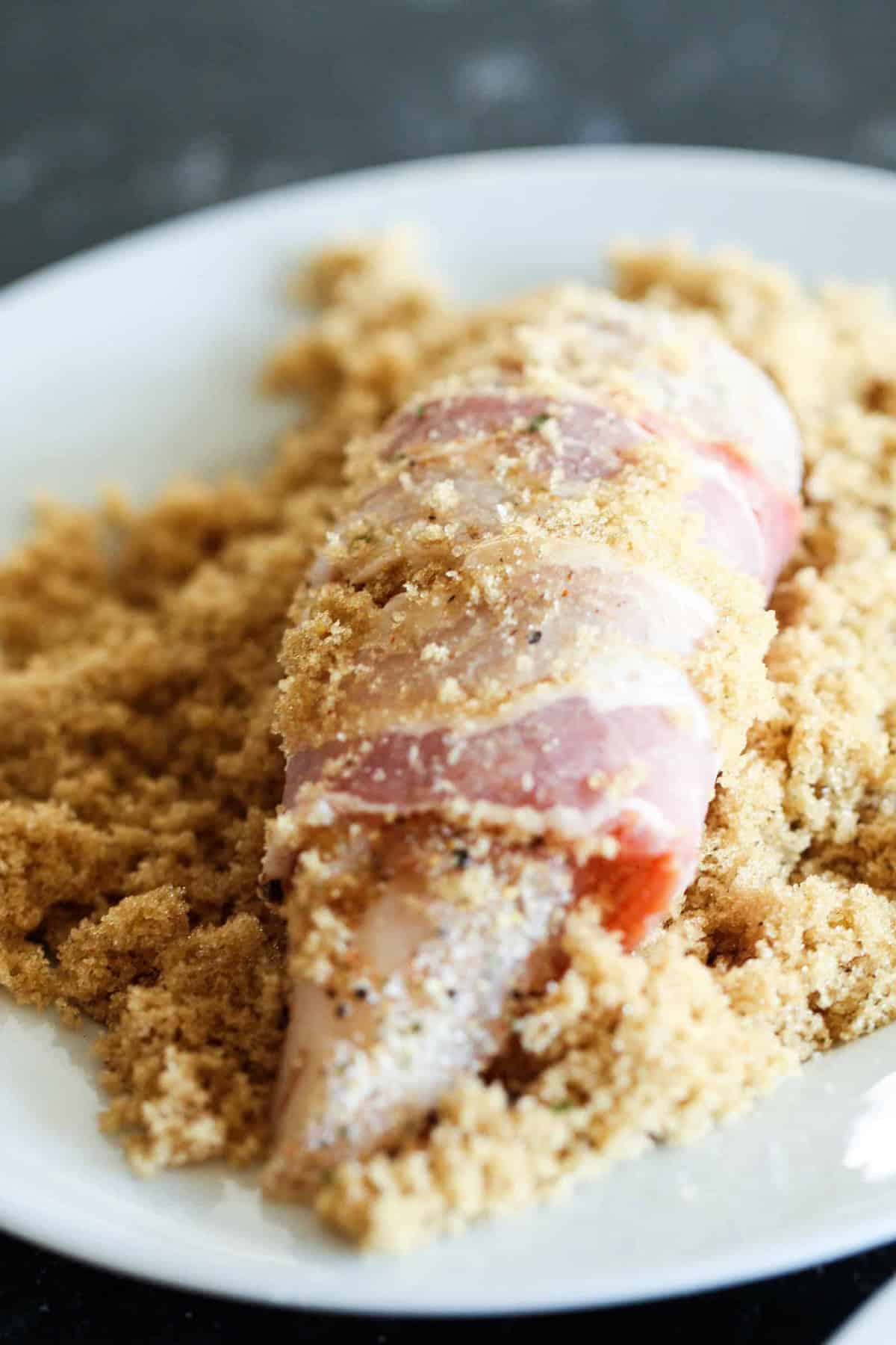 Raw chicken breast wrapped in bacon, coated with brown sugar, placed on a white plate with additional brown sugar around it.