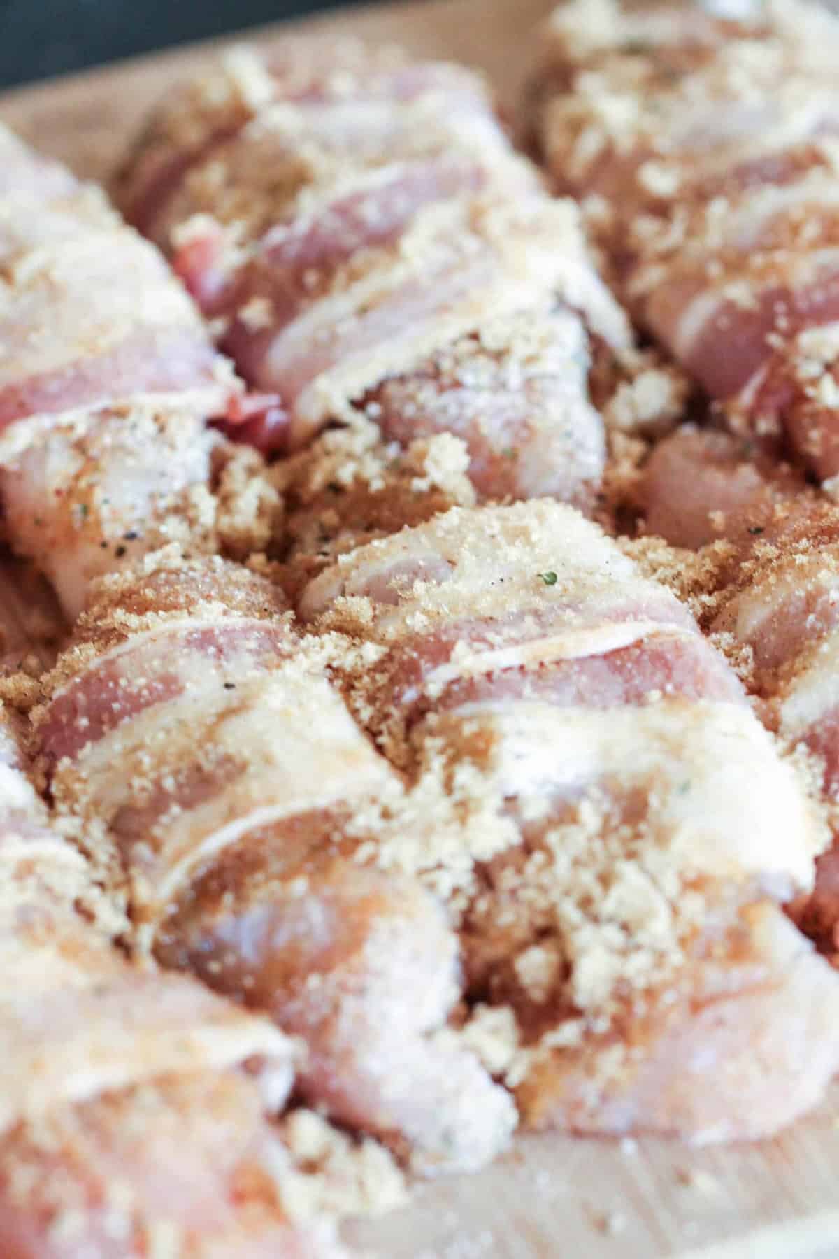 Close-up of chicken pieces wrapped in bacon, topped with bread crumbs, placed on a wooden surface.