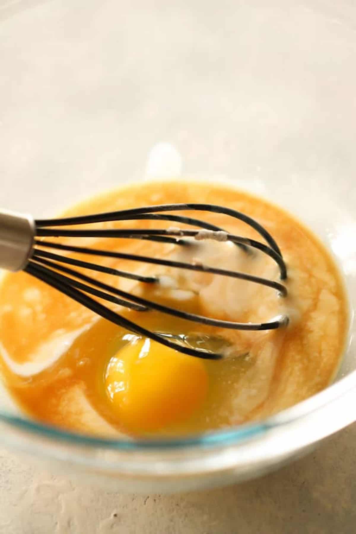 A whisk in a glass bowl with an egg and other wet baking ingredients, partially mixed.