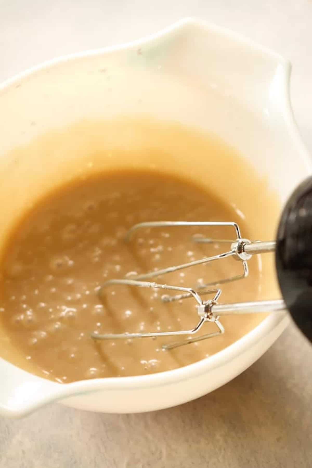 A mixing bowl with light brown batter being mixed by an electric hand mixer.