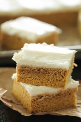 Two frosted pumpkin bars are stacked on a piece of wax paper, with another bar in the background on a tray. Each bar has a layer of creamy frosting on top of a dense pumpkin base.