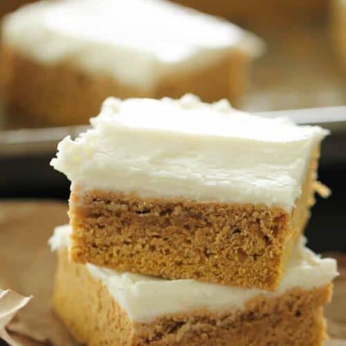 Two frosted pumpkin bars are stacked on a piece of wax paper, with another bar in the background on a tray. Each bar has a layer of creamy frosting on top of a dense pumpkin base.