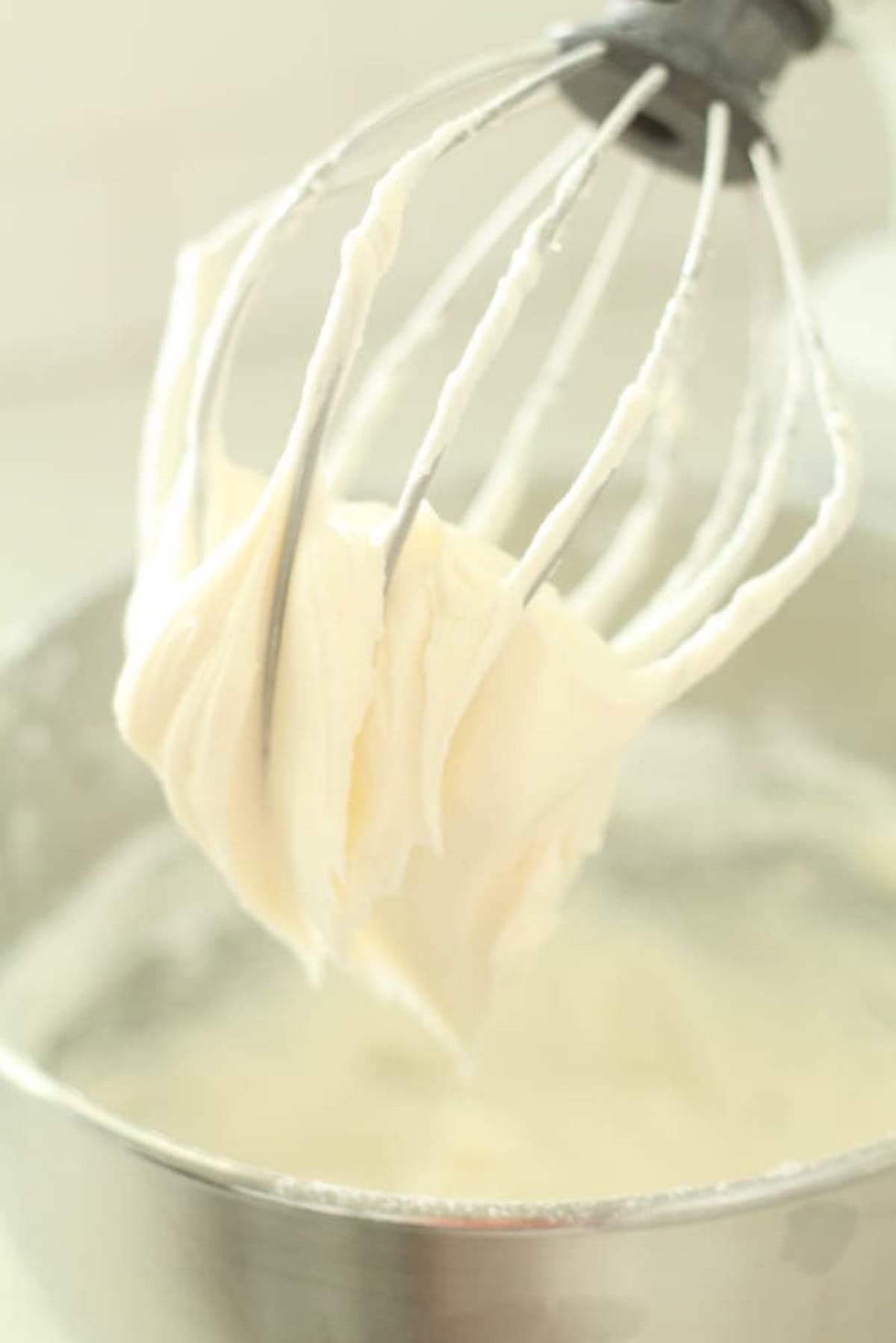 A metal whisk attachment covered in freshly whipped cream rests above a stainless steel mixing bowl.