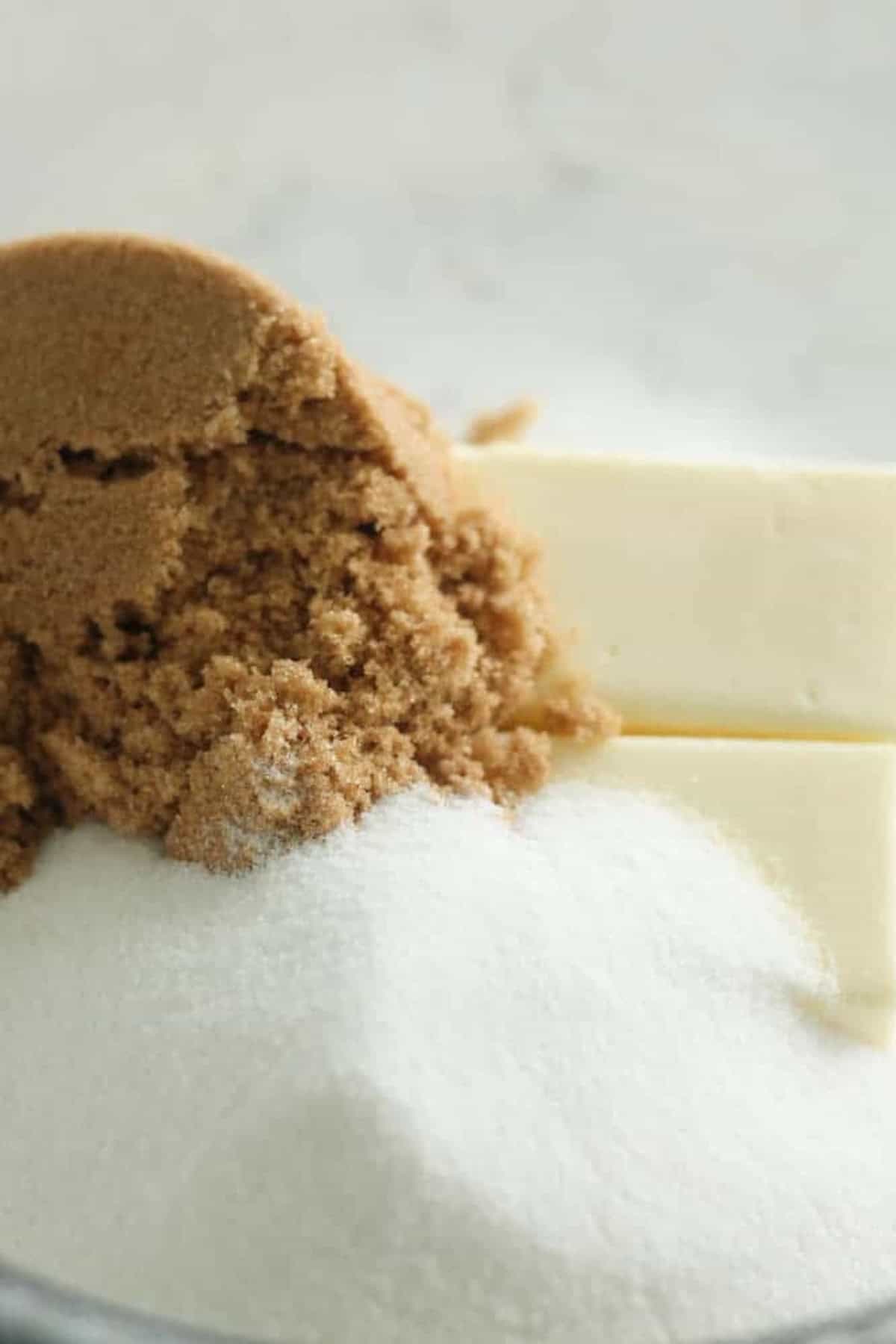 Close-up image showing a pile of granulated sugar, two sticks of butter, and a heap of brown sugar in a mixing bowl.