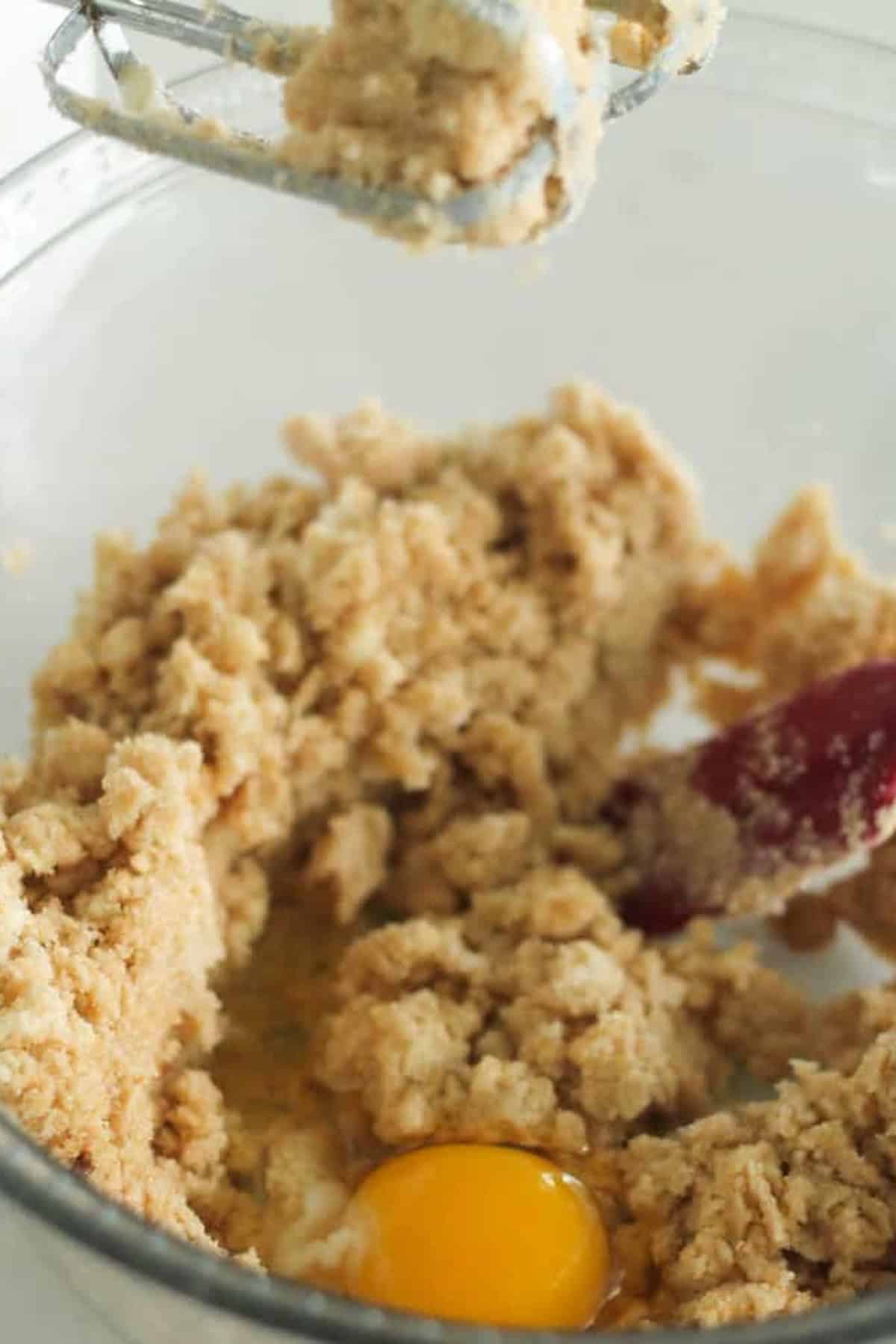 A mixing bowl containing a partially mixed dough with a visible egg and a red spatula. The edge of a hand mixer is seen above the bowl.