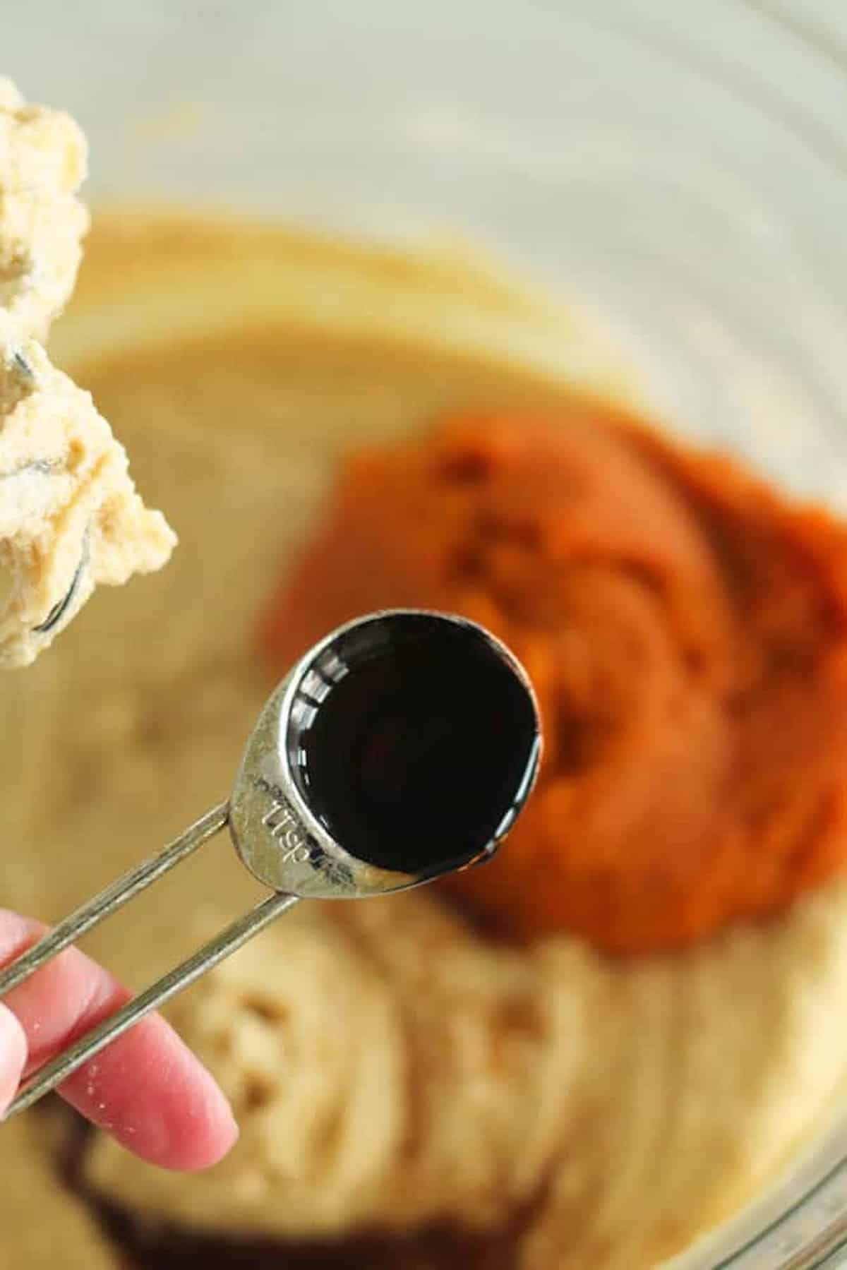 A person holds a teaspoon with a dark liquid over a mixing bowl containing batter and pumpkin puree.