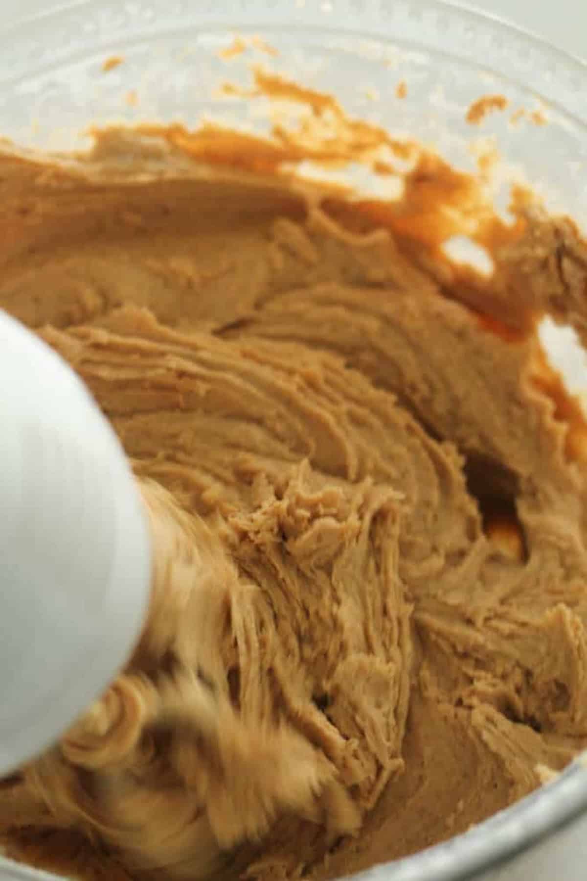 A close-up of light brown cookie dough being mixed by an electric mixer in a clear bowl. The mixture is thick and part of the mixer attachment is visible.