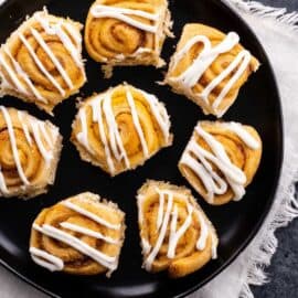 A black plate with six cinnamon rolls topped with white icing, displayed on a textured white and gray surface.