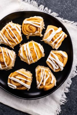 A black plate with six cinnamon rolls topped with white icing, displayed on a textured white and gray surface.