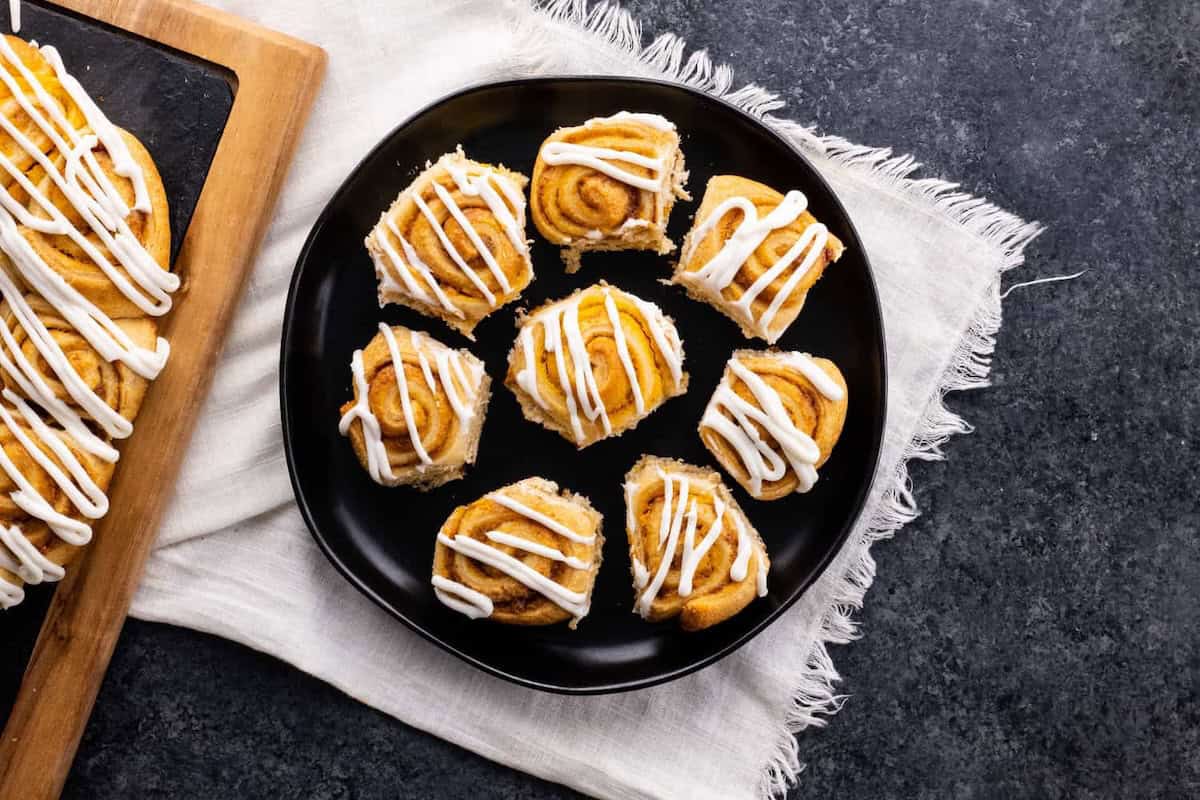 A black plate with seven small cinnamon rolls topped with white icing is placed on a white cloth. A wooden tray with more cinnamon rolls is partially visible on the left.