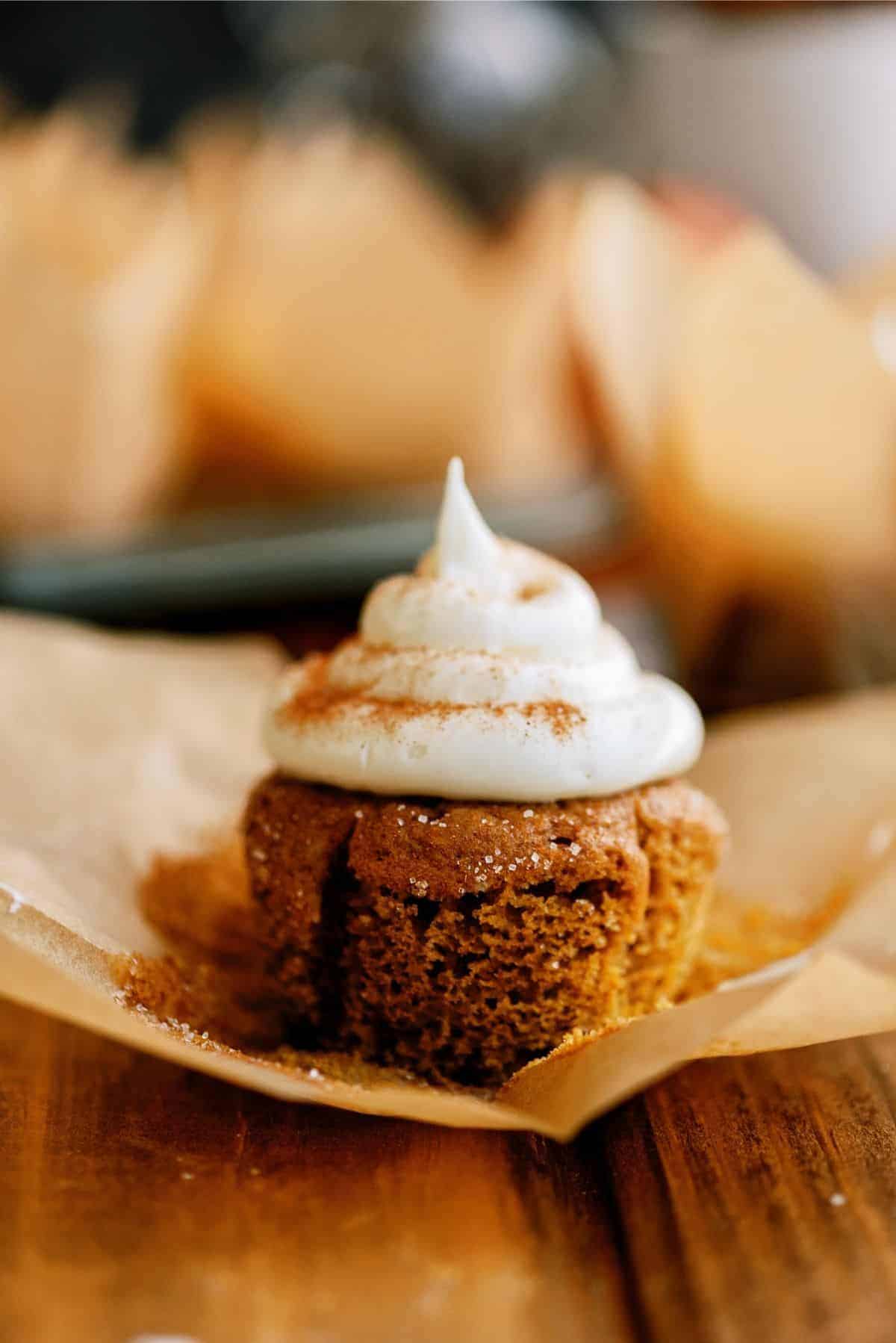 A cupcake with brown cake and white frosting, lightly dusted with cinnamon, sits on a piece of parchment paper on a wooden table.