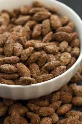 A white bowl filled with caramelized almonds, with additional almonds spilling out around the bowl.