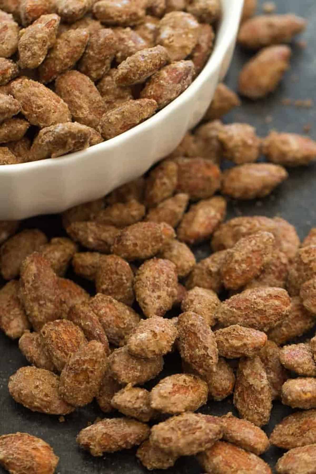 A white bowl filled with seasoned, roasted almonds is tipped over, with some of the almonds spilling out onto a dark surface.