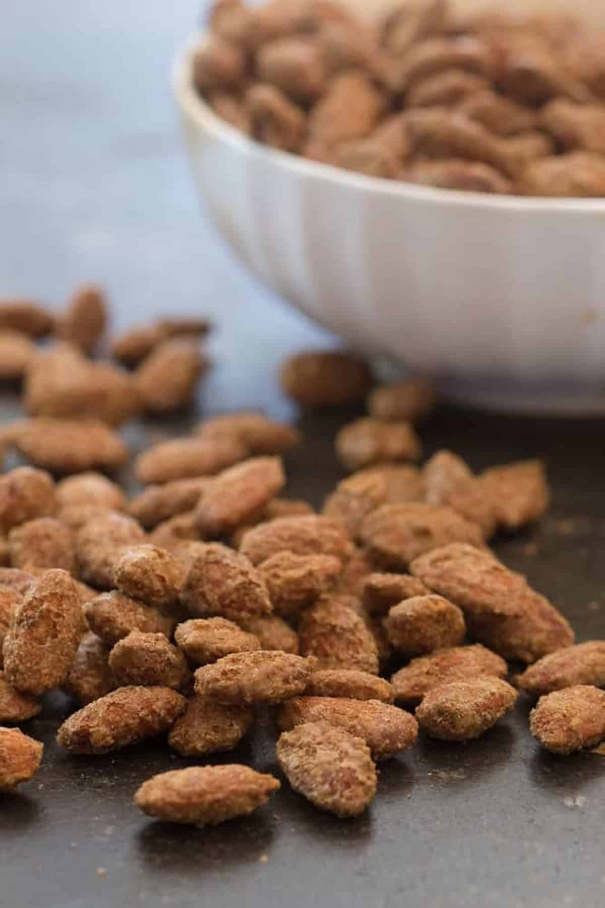 Close-up of sugar-coated almonds scattered on a dark surface, with a white bowl filled with more almonds in the background.