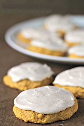 A plate of frosted cookies is in the background, with three frosted cookies in focus in the foreground on a dark surface.