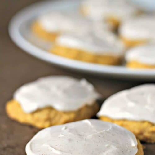 A plate of frosted cookies is in the background, with three frosted cookies in focus in the foreground on a dark surface.