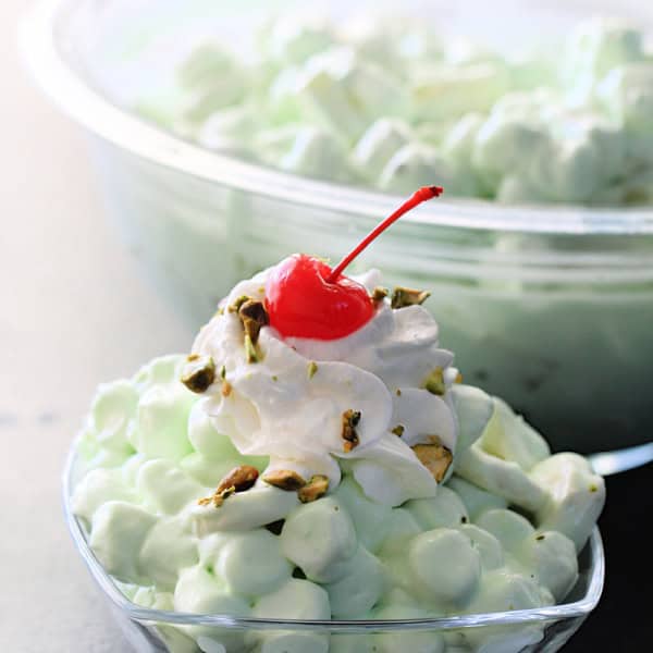 A close-up of a bowl of green pistachio salad topped with whipped cream, a maraschino cherry, and garnished with chopped nuts, with a larger bowl of the same salad in the background.