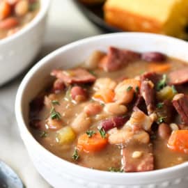 A bowl of hearty bean soup with diced vegetables and chunks of meat, garnished with herbs, served next to slices of cornbread.