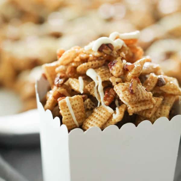A close-up of a white container filled with caramel-coated snack mix, featuring cereal pieces and pretzels, drizzled with a white icing.