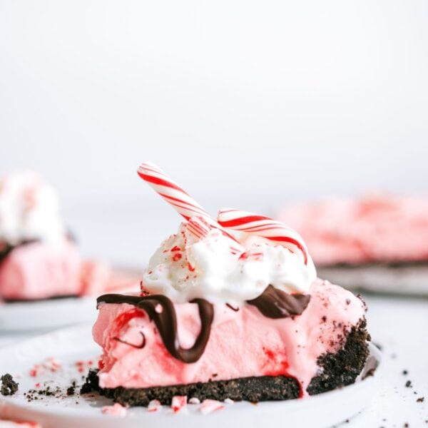 A slice of pink peppermint pie with a chocolate crust, topped with whipped cream, chocolate drizzle, and a peppermint stick, on a white plate.