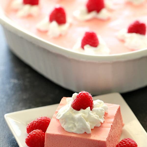 A square piece of pink gelatin dessert topped with whipped cream and raspberries sits on a small plate. A larger dish of the same dessert is seen in the background.