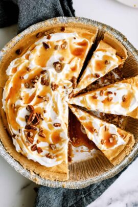 A pie with whipped cream topping, caramel drizzle, and pecan pieces, partially sliced and placed in a foil pie dish on a dark cloth.