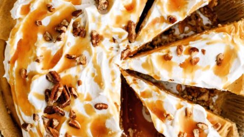 A pie with whipped cream topping, caramel drizzle, and pecan pieces, partially sliced and placed in a foil pie dish on a dark cloth.