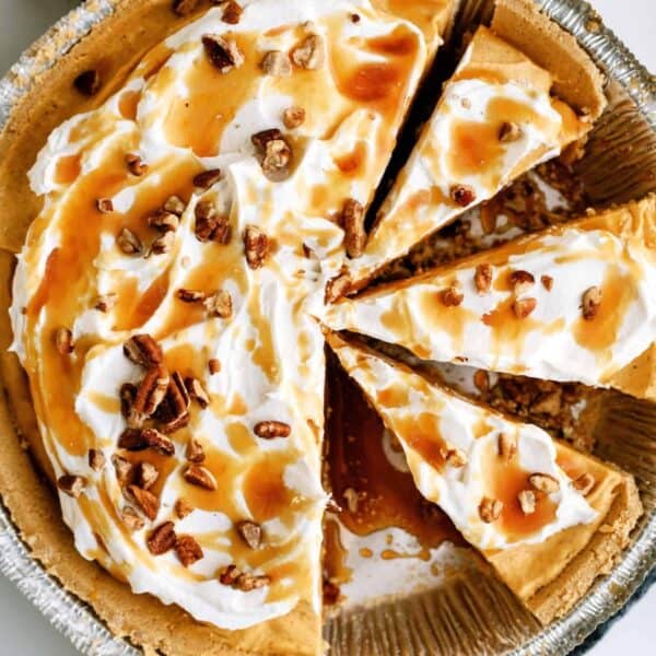 A pie with whipped cream topping, caramel drizzle, and pecan pieces, partially sliced and placed in a foil pie dish on a dark cloth.