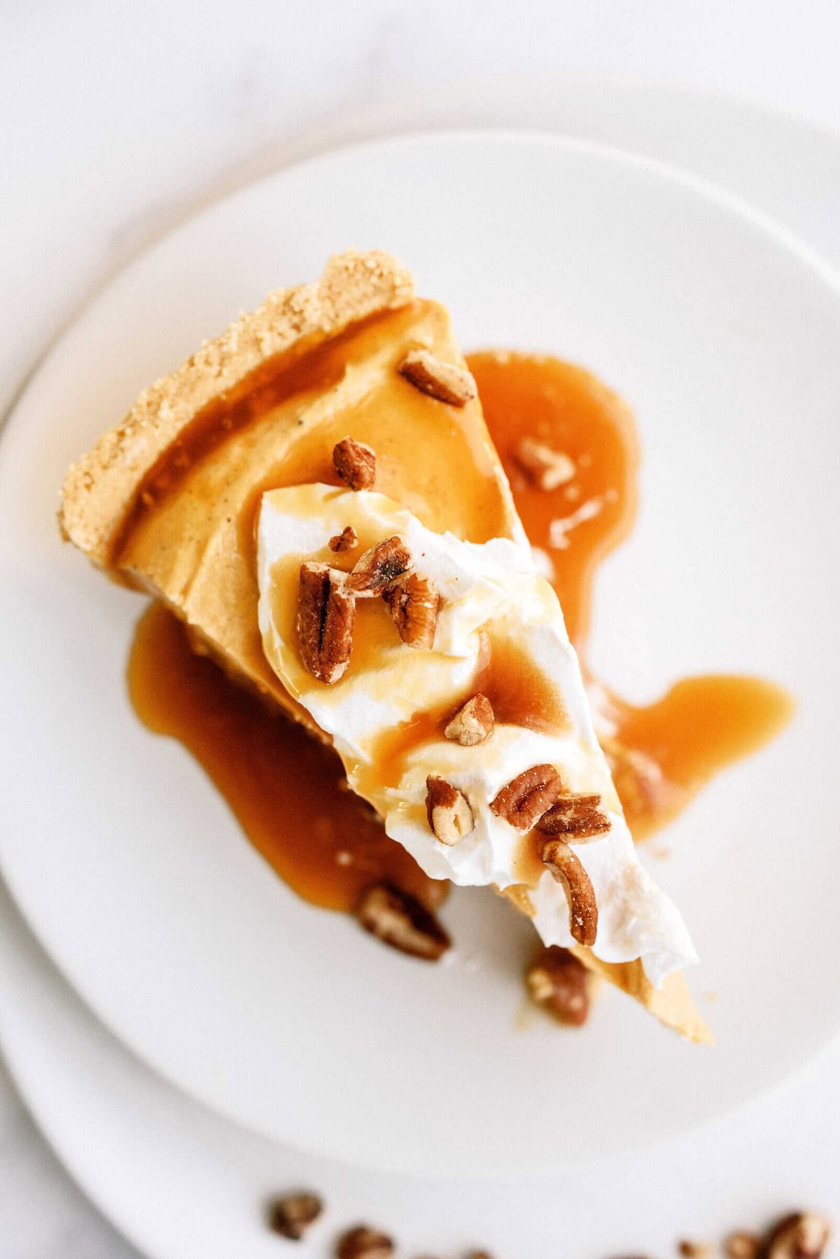 A slice of pie with a golden-brown crust, topped with whipped cream, chopped pecans, and drizzled with caramel sauce, on a white plate.