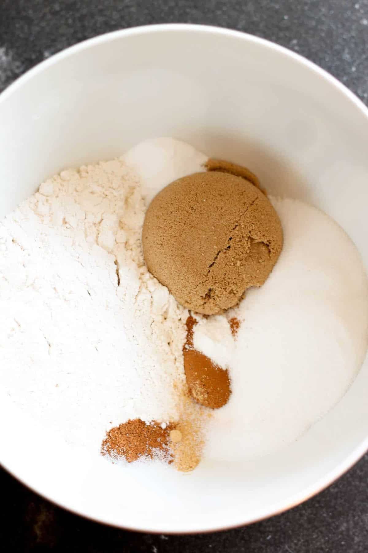 A white bowl containing flour, white sugar, brown sugar, cinnamon, and nutmeg on a dark countertop.