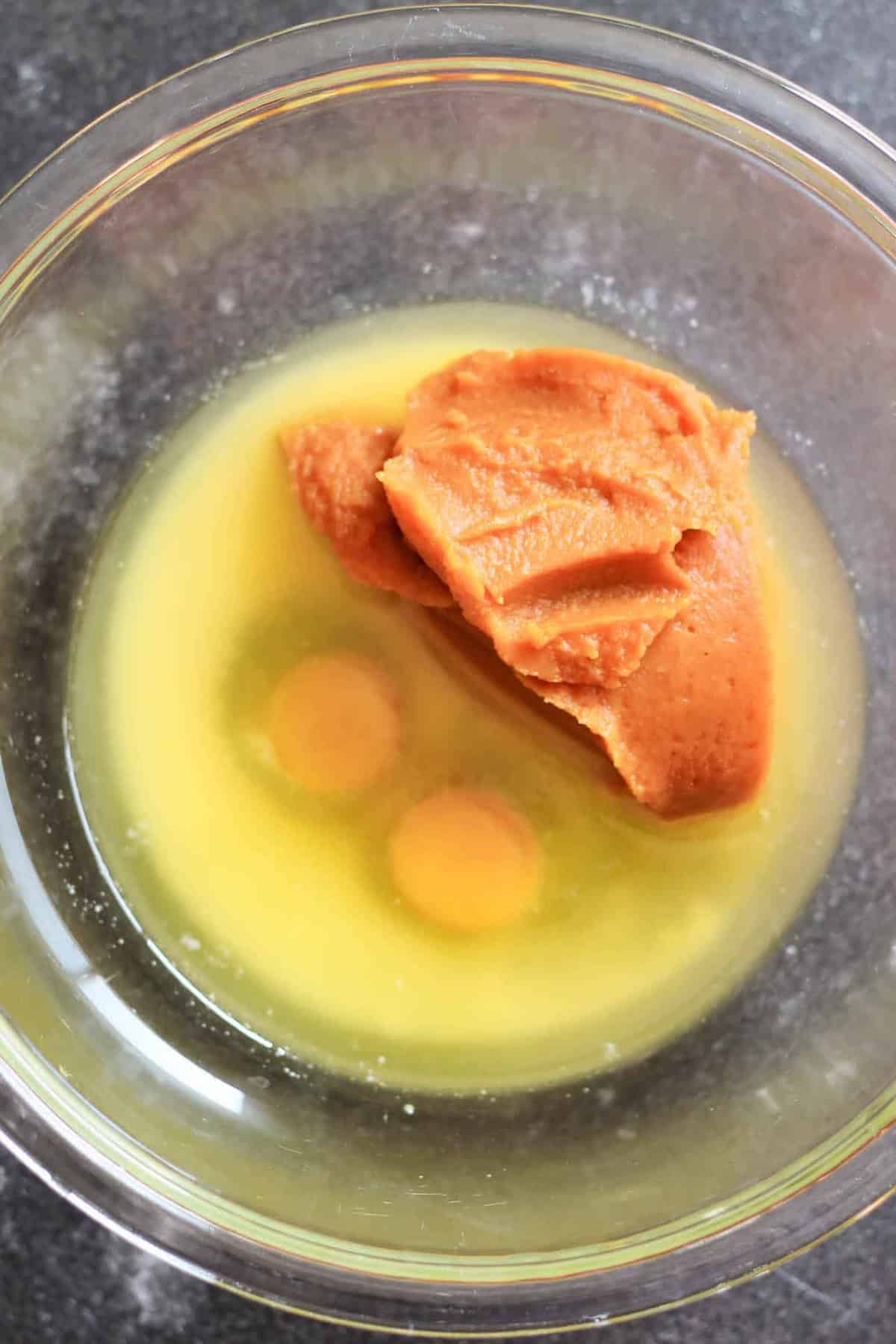 A glass bowl containing two raw eggs, melted butter, and a large dollop of peanut butter.