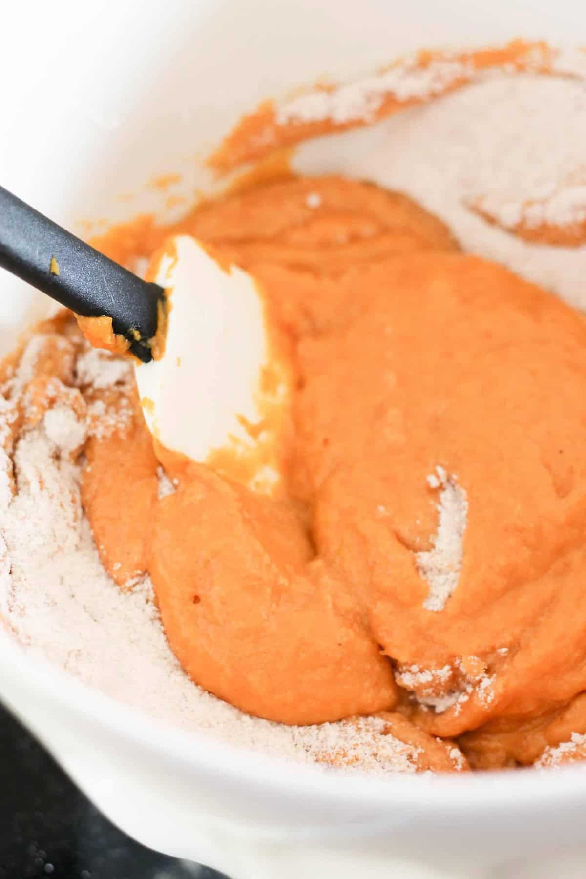 A white bowl containing a pumpkin batter being mixed with a black spatula. Some flour is visible on the sides of the bowl.