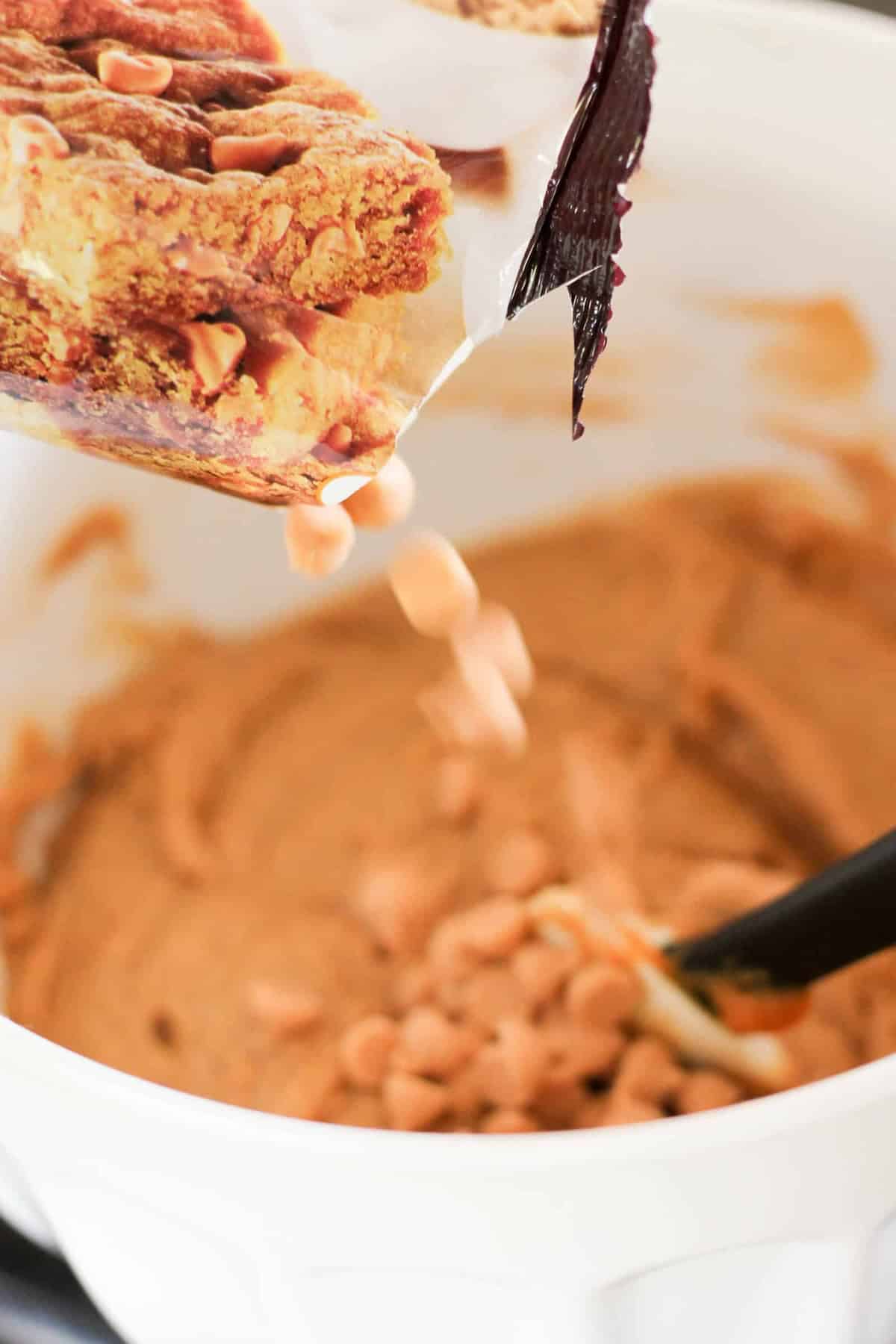 Pouring butterscotch chips from a bag into a bowl of cookie dough with a black spatula.