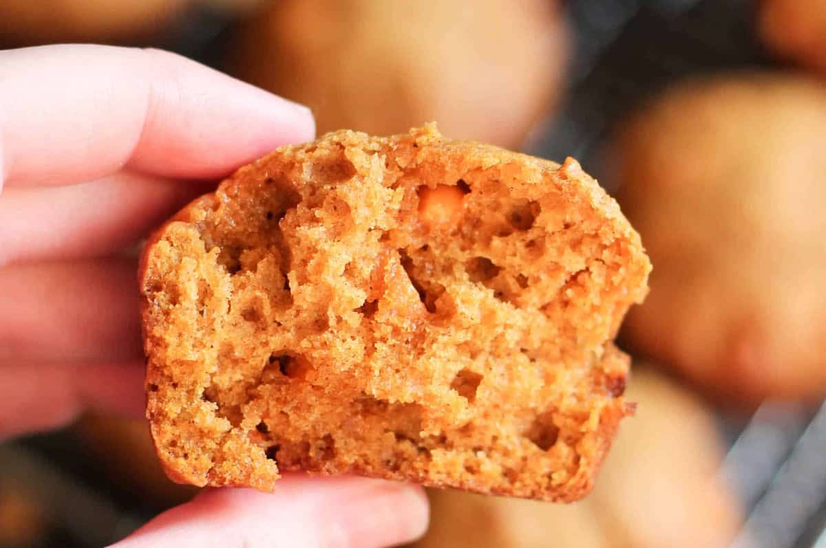 A hand holding a half-eaten baked muffin, revealing a moist, crumbly texture and orange bits inside.