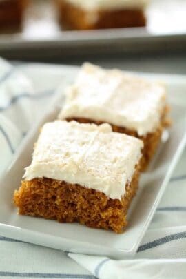 Two rectangular slices of frosted pumpkin bars are placed on a white dish, atop a striped cloth.