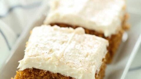 Two rectangular slices of frosted pumpkin bars are placed on a white dish, atop a striped cloth.