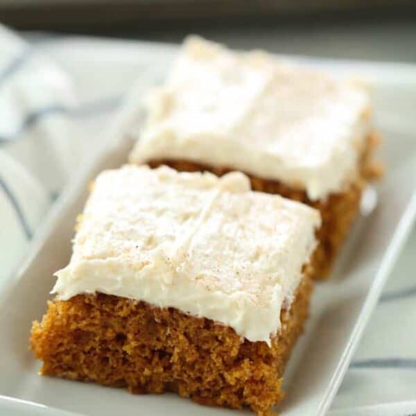 Two rectangular slices of frosted pumpkin bars are placed on a white dish, atop a striped cloth.