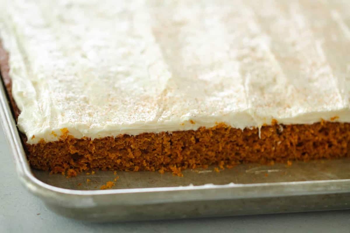 A close-up of a frosted rectangular sheet cake on a baking tray with a piece missing from one corner.