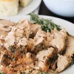 A plate of sliced herb-crusted turkey garnished with rosemary, alongside a bowl of gravy and dinner rolls.