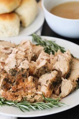 A plate of sliced herb-crusted turkey garnished with rosemary, alongside a bowl of gravy and dinner rolls.