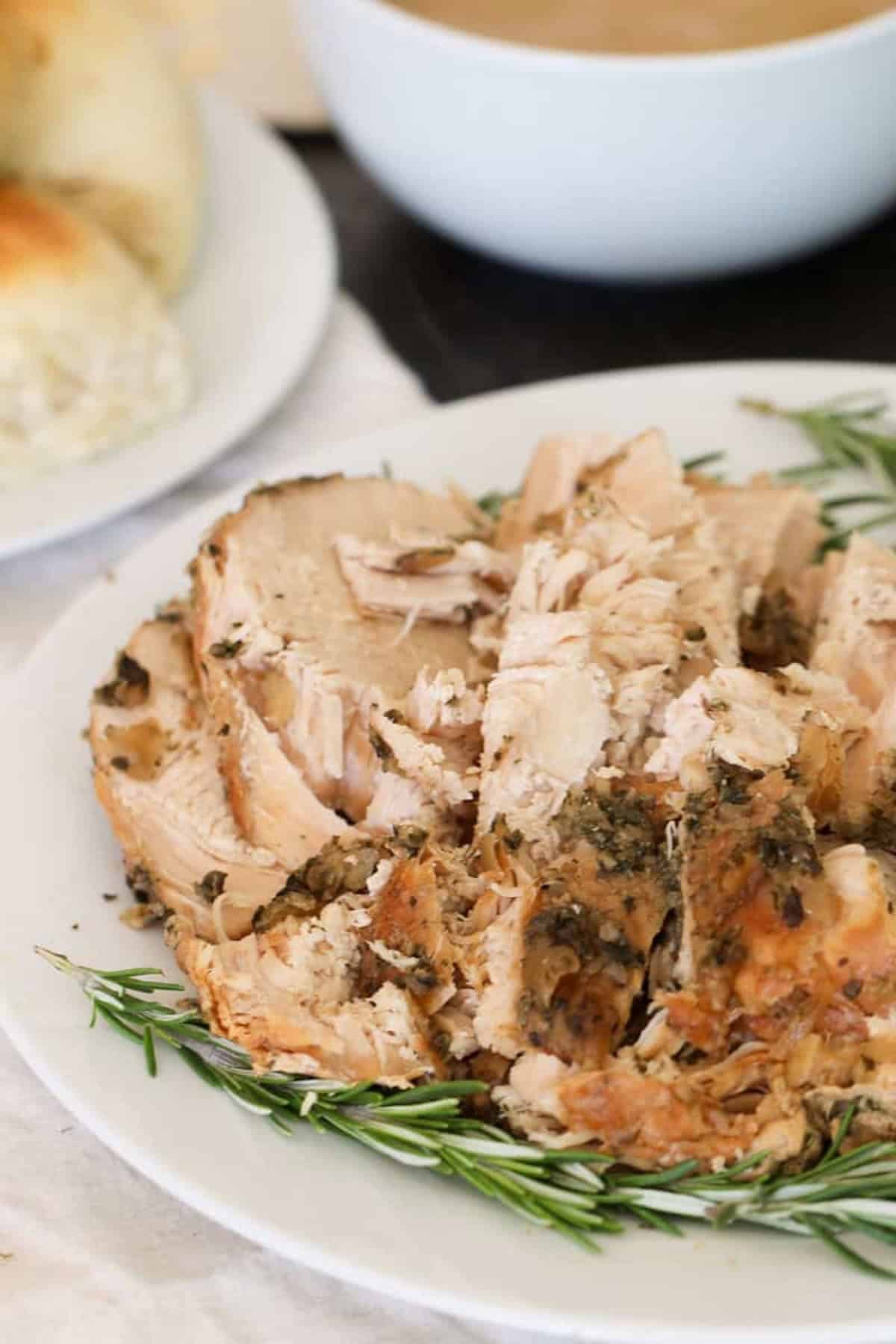 Slices of seasoned roast turkey on a white plate garnished with rosemary, with bread rolls and a bowl of gravy in the background.