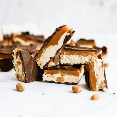 Several chocolate-covered caramel and nougat bars with peanuts, some cut in half, are displayed on a white surface.