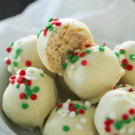 A bowl of white chocolate truffles decorated with red, white, and green sprinkles. One truffle has a bite taken out, revealing its crumbly interior.