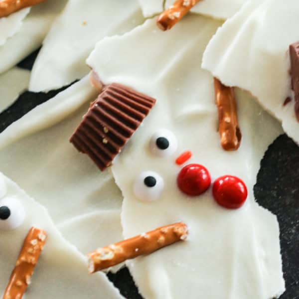 White chocolate bark decorated with candy eyes, red candies for buttons, mini peanut butter cup hats, and pretzel sticks for arms, resembling snowmen.