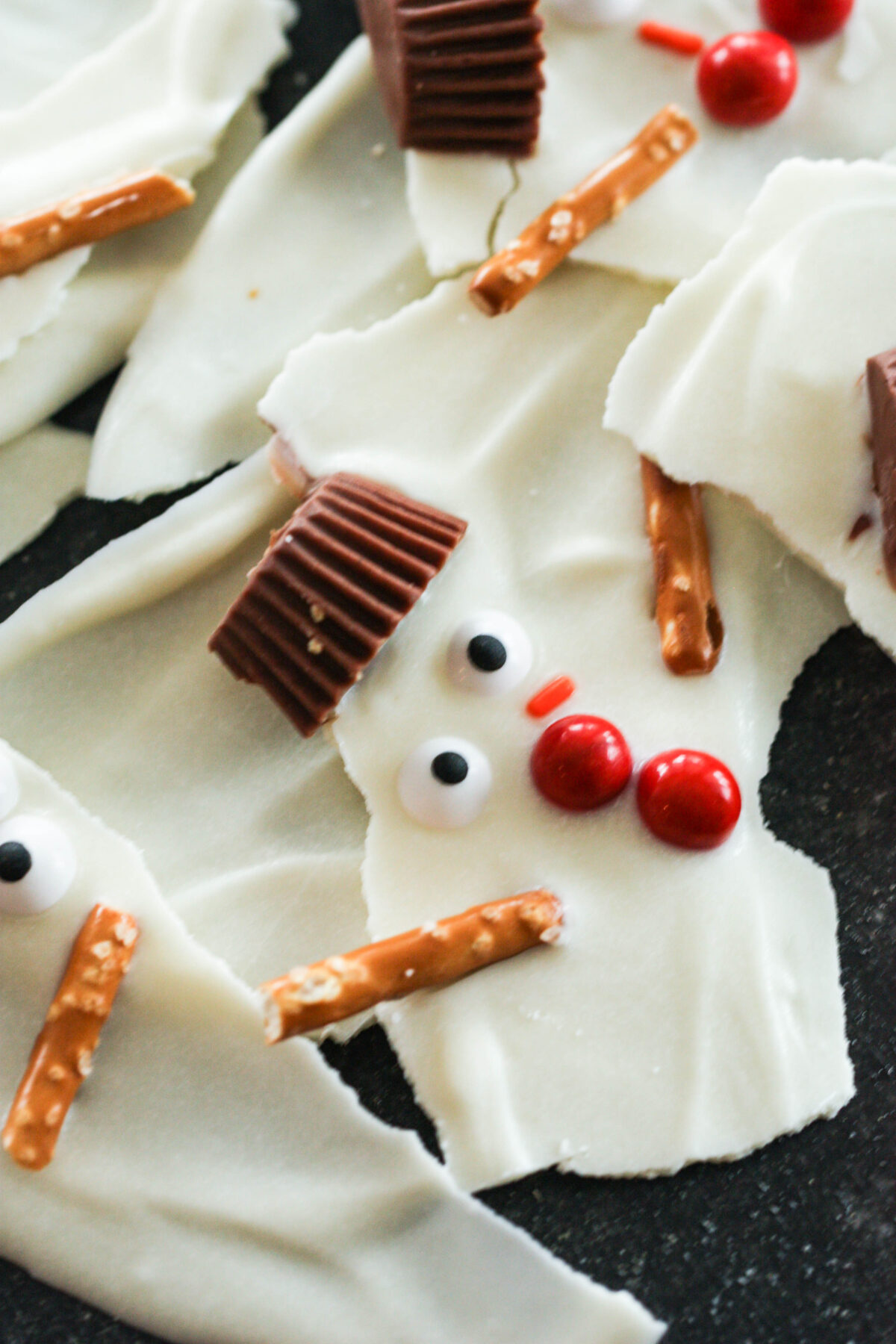 White chocolate bark decorated with candy eyes, red candies for buttons, mini peanut butter cup hats, and pretzel sticks for arms, resembling snowmen.