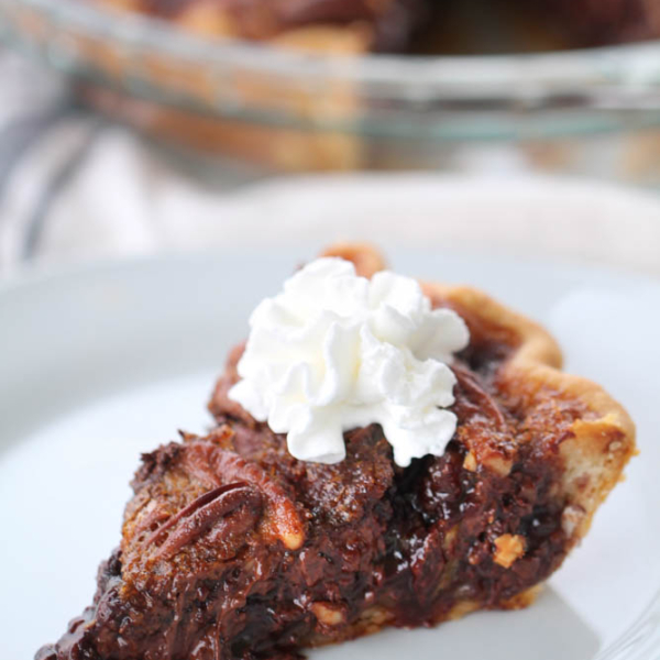 Slice of chocolate pecan pie topped with whipped cream on a white plate.