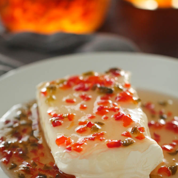 A block of cream cheese covered in a sauce with chopped red and green peppers, served on a white plate.